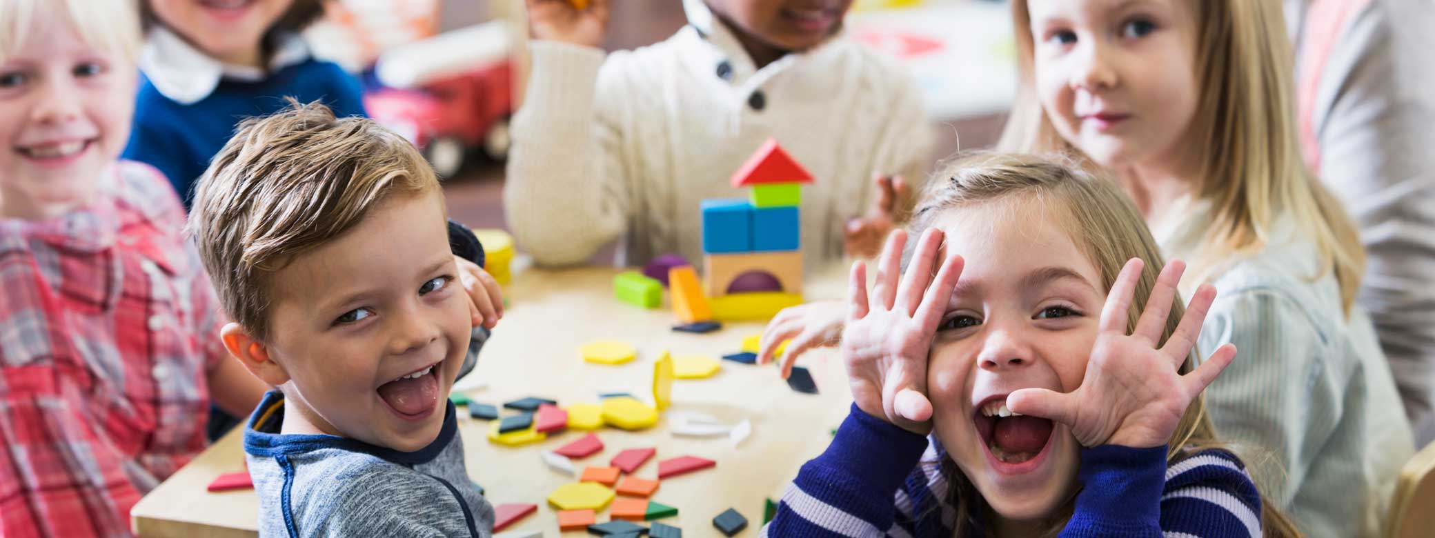 Kinder sitzen am Tisch mit Bauklötzen und schauen freudig nach vorne